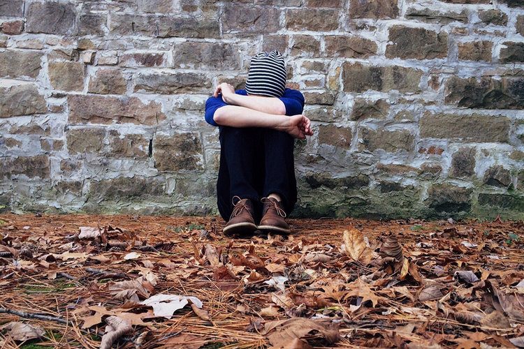 Eine Person sitzt auf Laub vor einer Steinmauer, die Beine angezogen, den Kopf gesenkt und versteckt in seinen Armen - Copyright: Bastian Modrow